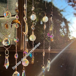 Peut inclure: Plusieurs attrape-soleil en cristal suspendus devant une fen&ecirc;tre. Les d&eacute;corations comprennent des cristaux clairs et facett&eacute;s, des perles iris&eacute;es et des &eacute;l&eacute;ments m&eacute;talliques dor&eacute;s. Les motifs incluent des soleils et des lunes, ainsi que divers pendentifs en cristal. Le nom de la boutique Etsy "elena monroe boutique" est visible.