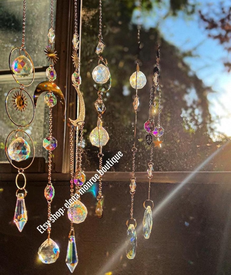 Peut inclure: Plusieurs attrape-soleil en cristal suspendus devant une fen&ecirc;tre. Les d&eacute;corations comprennent des cristaux clairs et facett&eacute;s, des perles iris&eacute;es et des &eacute;l&eacute;ments m&eacute;talliques dor&eacute;s. Les motifs incluent des soleils et des lunes, ainsi que divers pendentifs en cristal. Le nom de la boutique Etsy "elena monroe boutique" est visible.