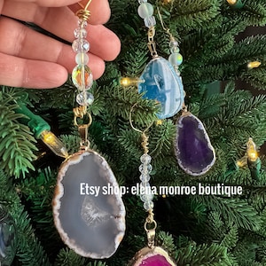 May include: Christmas tree ornaments with natural agate slices in shades of gray, purple, and pink, trimmed with gold. Each ornament is suspended by a gold wire and adorned with clear and iridescent beads. The text "Etsy shop: elena monroe boutique" is visible.