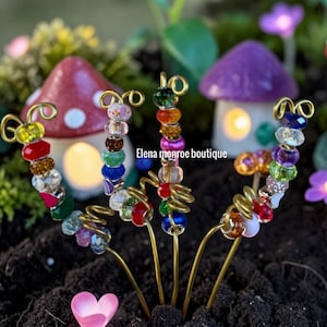 Può includere: Primo piano di paletti da giardino con perline colorate con accenti di filo d'oro. I paletti presentano una varietà di perline dai colori gioiello e sono disposti nel terreno con case a fungo in miniatura sullo sfondo. Il testo "Elena monroe boutique" è visibile.