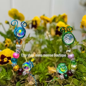 May include: Two decorative garden stakes with colorful beads and iridescent crystal accents. The stakes have a wire design with a spiral top, and are set against a backdrop of yellow flowers and green foliage. The text "Etsy shop: elena monroe Boutique" is visible.