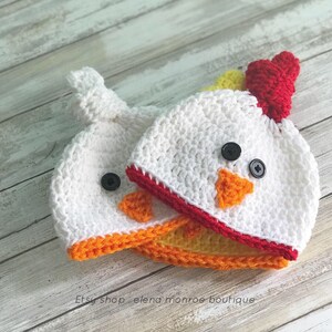 K&ouml;nnte beinhalten: Zwei geh&auml;kelte Baby-M&uuml;tzen in Form eines K&uuml;ken. Die M&uuml;tzen sind wei&szlig; mit orangefarbenem und gelbem Schnabel, rotem Kamm und schwarzen Knopfaugen. Etsy-Shop elena monroe boutique.