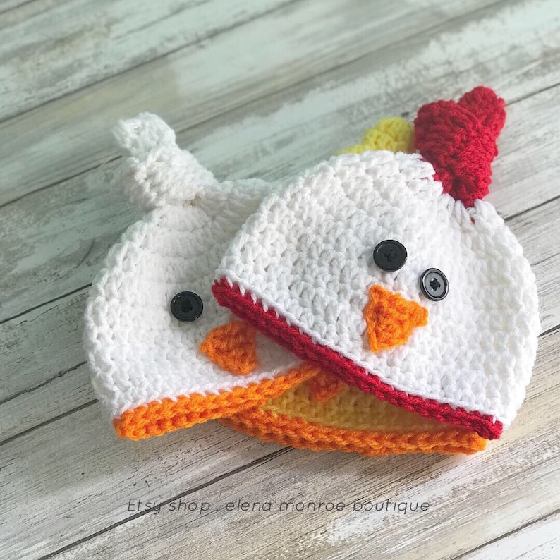 K&ouml;nnte beinhalten: Zwei geh&auml;kelte Baby-M&uuml;tzen in Form eines K&uuml;ken. Die M&uuml;tzen sind wei&szlig; mit orangefarbenem und gelbem Schnabel, rotem Kamm und schwarzen Knopfaugen. Etsy-Shop elena monroe boutique.