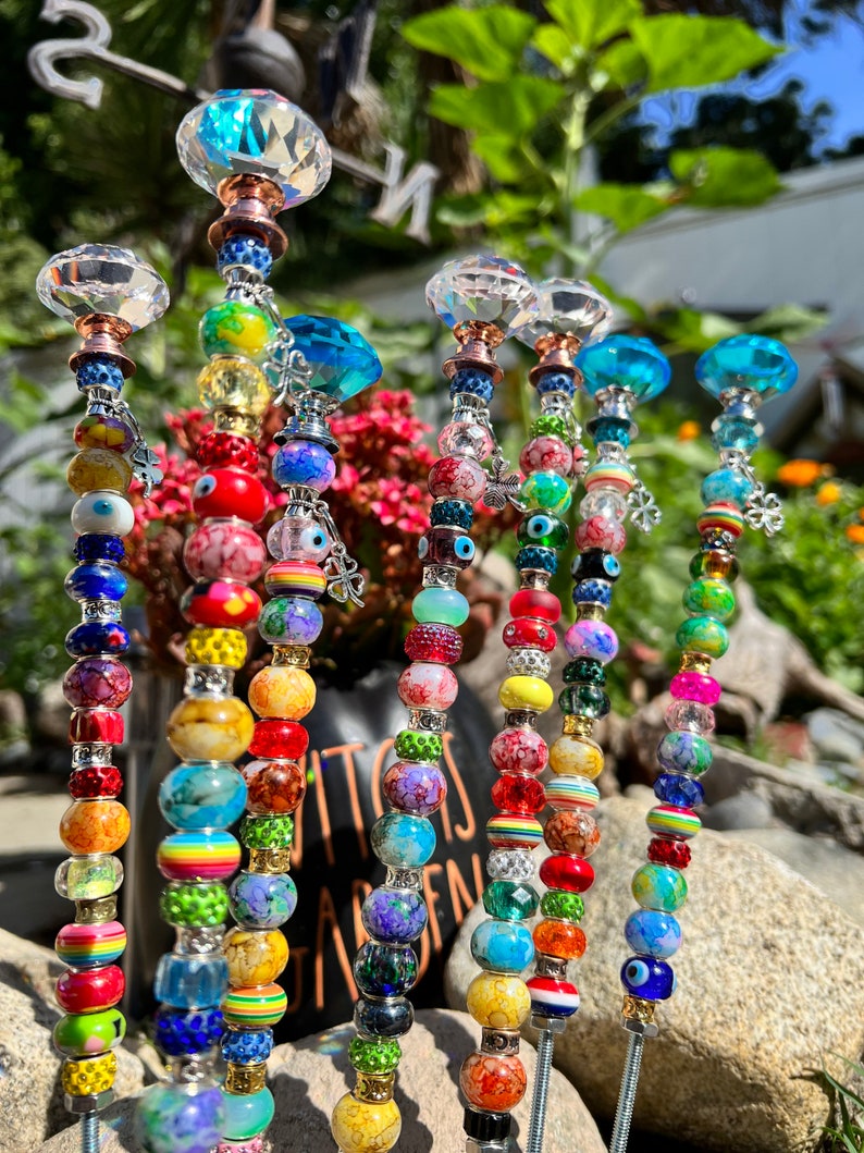 May include: A set of colorful beaded garden stakes with crystal tops. Each stake features a variety of beads in different colors, patterns, and textures, including glass, wood, and metal. The stakes are designed to add a whimsical touch to any garden.