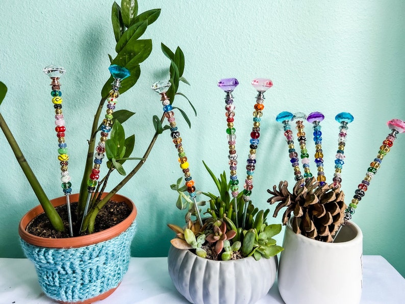 May include: A set of colorful beaded plant stakes with crystal tops. The stakes are inserted into three different potted plants, including a succulent and a leafy plant.