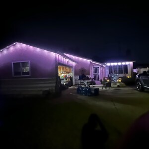 K&ouml;nnte beinhalten: Ein Haus von au&szlig;en bei Nacht, beleuchtet mit lila und wei&szlig;en Lichterketten. Die Dachlinie und die Fenster sind mit Lichtern umrandet, was eine festliche Atmosph&auml;re schafft. Ein Auto steht in der Einfahrt.