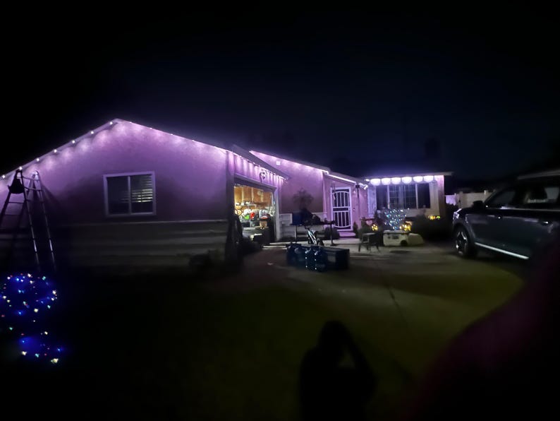 K&ouml;nnte beinhalten: Ein Haus von au&szlig;en bei Nacht, beleuchtet mit lila und wei&szlig;en Lichterketten. Die Dachlinie und die Fenster sind mit Lichtern umrandet, was eine festliche Atmosph&auml;re schafft. Ein Auto steht in der Einfahrt.