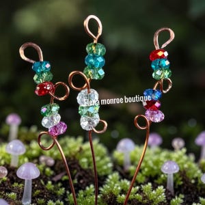 May include: Three copper wire plant stakes, each decorated with vibrant, faceted beads in red, green, blue, and clear. The stakes are set against a backdrop of green moss and small purple mushroom figurines.