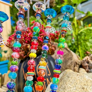 May include: Six colorful beaded garden stakes with crystal tops. Each stake features a variety of beads in different colors, patterns, and textures, including rainbow, green, blue, and white beads. The stakes are topped with clear crystal finials.