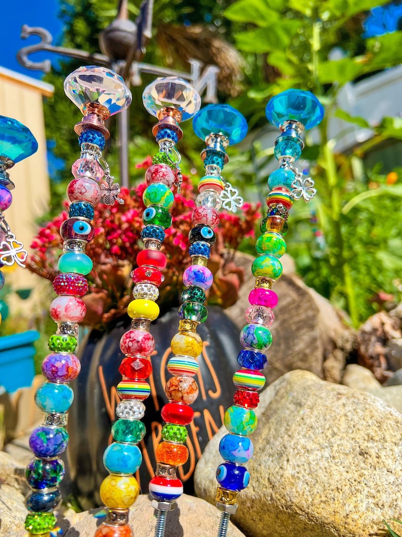 May include: Six colorful beaded garden stakes with crystal tops. Each stake features a variety of beads in different colors, patterns, and textures, including rainbow, green, blue, and white beads. The stakes are topped with clear crystal finials.