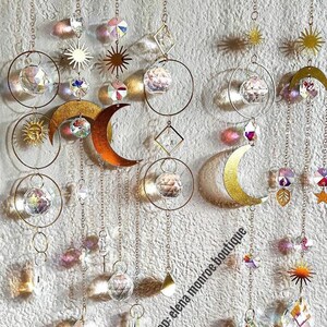 Peut inclure: Suncatchers d&eacute;coratifs suspendus avec des soleils, des lunes et des formes g&eacute;om&eacute;triques en m&eacute;tal dor&eacute;. Les capteurs de soleil pr&eacute;sentent des boules et des prismes en cristal transparent, suspendus par de fines cha&icirc;nes. Le nom de la boutique Etsy "elena monroe boutique" est visible.