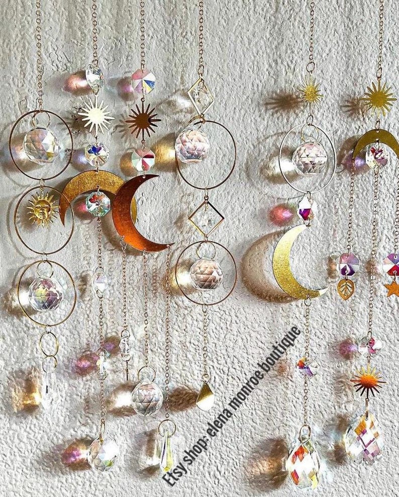 Peut inclure: Suncatchers d&eacute;coratifs suspendus avec des soleils, des lunes et des formes g&eacute;om&eacute;triques en m&eacute;tal dor&eacute;. Les capteurs de soleil pr&eacute;sentent des boules et des prismes en cristal transparent, suspendus par de fines cha&icirc;nes. Le nom de la boutique Etsy "elena monroe boutique" est visible.