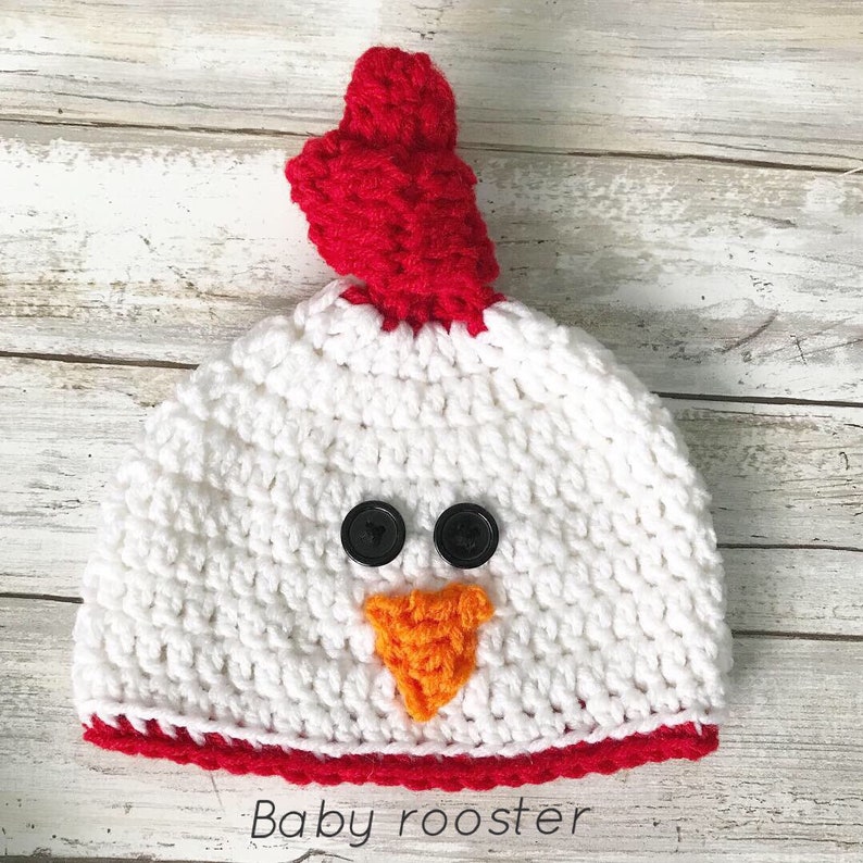 K&ouml;nnte beinhalten: Wei&szlig;e geh&auml;kelte Baby-M&uuml;tze mit rotem Kamm, orangem Schnabel und schwarzen Knopfaugen. Die M&uuml;tze hat einen roten Saum am unteren Rand. "Baby rooster" steht unter der M&uuml;tze.