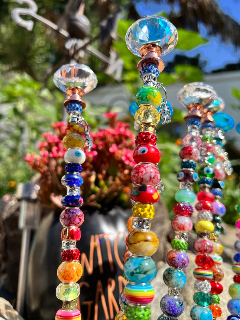 May include: A set of colorful beaded garden stakes with crystal tops. The beads are various colors, including red, blue, green, yellow, and orange. The stakes are made of metal and have a silver finish. The crystal tops are clear and have a faceted design.