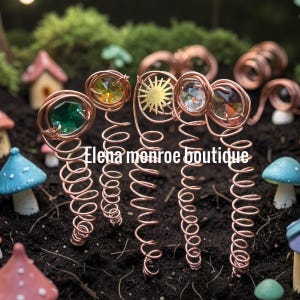 May include: Copper wire hair accessories with colorful crystal and sunburst accents. The accessories are arranged on a dark surface with miniature fairy garden elements in the background. The text "Elena monroe boutique" is visible.