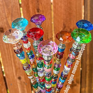May include: A collection of decorative beaded wands with faceted crystal toppers in various colors, including clear, blue, purple, red, orange, and green. The wands are adorned with an assortment of colorful beads and metallic accents. The Etsy shop name "elena monroe boutique" is visible.