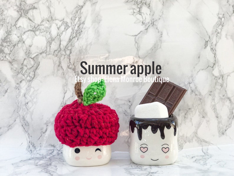 May include: Two white ceramic mugs shaped like marshmallows with smiling faces. One mug has a red crocheted apple hat on top and the other mug has a chocolate bar and a white marshmallow on top. The mugs are on a white marble background. The text "Summer apple" and "Etsy shop Elena Monroe Boutique" is written above the mugs.