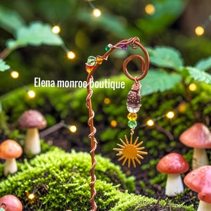 May include: A copper-colored metal garden stake with a sun charm and colorful beads. The stake has a spiral design and the text "Elena monroe boutique". The stake is placed in a mossy garden setting with mushrooms.