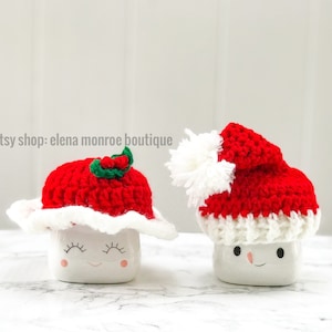May include: Two white ceramic mugs with happy faces wearing red and white crocheted Santa hats. One hat has a green holly sprig.