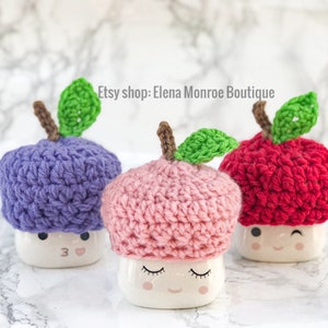 May include: Three crocheted apple-shaped mug toppers in purple, pink, and red. Each topper has a green crocheted leaf and sits on a white mug with a cartoon face.