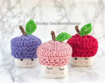 May include: Three crocheted apple-shaped mug toppers in purple, pink, and red. Each topper has a green crocheted leaf and sits on a white mug with a cartoon face.