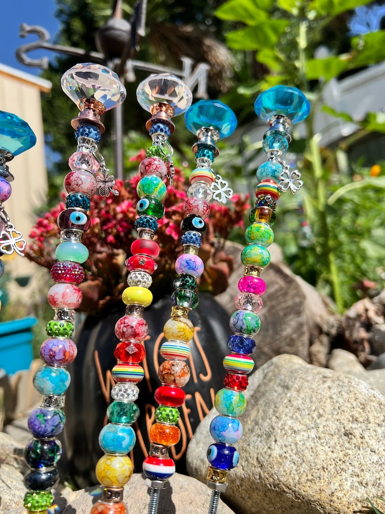 May include: Six colorful beaded garden stakes with crystal tops. Each stake features a variety of beads in different colors, patterns, and textures, including clear, blue, green, red, yellow, and pink beads. The stakes are topped with clear and blue crystal finials.