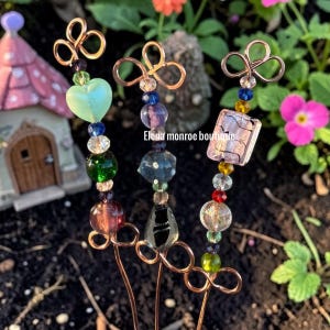 May include: Three decorative garden stakes with copper wire accents and colorful glass beads. Each stake features a unique design, including a heart, square, and round beads in shades of green, blue, purple, and gold. The text "Elena monroe boutique" is visible.