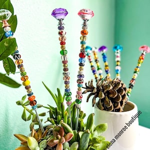 May include: Colorful beaded plant stakes with diamond-shaped tops in pink, purple, and clear. The stakes are inserted into a pot of succulents and a white ceramic pot with the text "Etsy shop: elena monroe boutique".