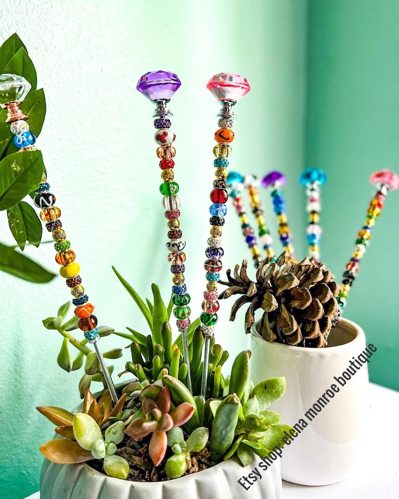 May include: Colorful beaded plant stakes with diamond-shaped tops in pink, purple, and clear. The stakes are inserted into a pot of succulents and a white ceramic pot with the text "Etsy shop: elena monroe boutique".