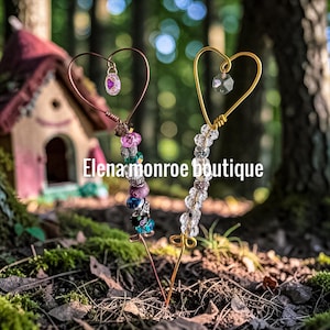 May include: Two decorative garden stakes featuring wire heart tops and colorful glass beads. One stake has a copper-toned wire with pink, blue, and purple beads, while the other has gold-toned wire with clear beads. Text reads Elena monroe boutique.