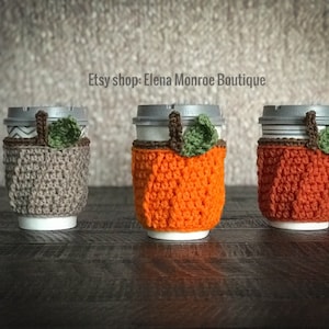 May include: Three crocheted pumpkin coffee cup cozies. One is brown, and two are orange. The cozies are on a dark wood surface. Etsy shop: Elena Monroe Boutique