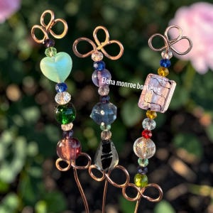 May include: Three decorative hair pins with copper wire accents and colorful beads. Each pin features a unique design, including a heart-shaped bead, a rectangular bead with a floral pattern, and various round and faceted beads. The text "Elena monroe boutique" is visible.