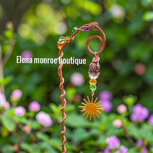 Pode incluir: Uma estaca de jardim de cobre artesanal com um pingente de sol, contas coloridas e um elemento decorativo em forma de sino. A estaca tem um design torcido e o texto "Elena monroe boutique".