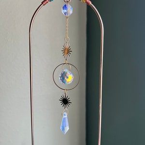 May include: A gold metal stand with a crystal suncatcher hanging from it. The suncatcher features a variety of crystal shapes, including a teardrop, a sphere, and a star. The stand is decorated with a beaded garland.