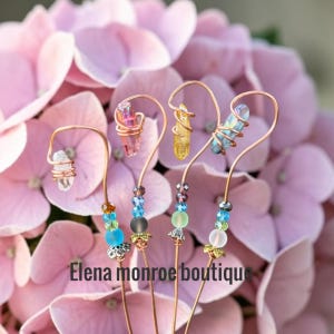 May include: Four copper wire plant stakes, each topped with a raw crystal point in clear, pink, yellow, or blue. The stems feature decorative glass and metal beads in shades of blue, green, and clear. The text Elena monroe boutique is visible.