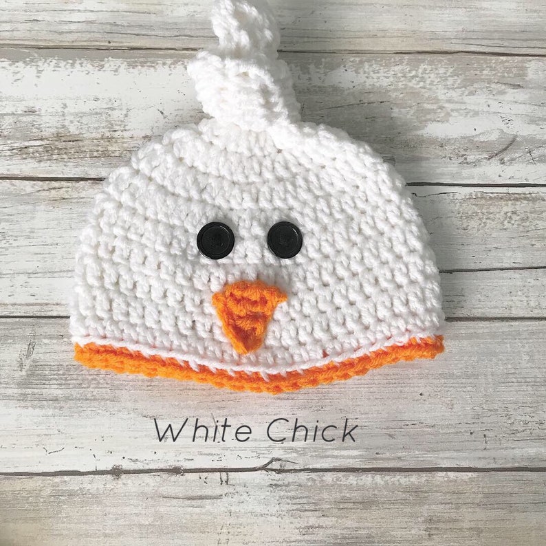 K&ouml;nnte beinhalten: Wei&szlig;e H&auml;kelm&uuml;tze f&uuml;r Babys mit orangem Saum und schwarzen Knopfaugen. Die M&uuml;tze ist in Form eines K&uuml;ken mit einem orangefarbenen Schnabel geformt.