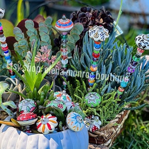 May include: A collection of colorful garden stakes with decorative ceramic tops and beaded stems. The stakes are placed in a pot of succulents, showcasing a variety of colors and textures. The ceramic tops feature hand-painted designs in red, white, green, and orange.