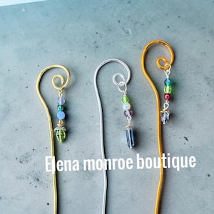 May include: Three metal bookmarks with curved tops, each featuring a dangling charm made of small colorful beads. The left bookmark is gold-toned, the center is silver-toned, and the right is gold-toned. The text Elena monroe boutique is visible.