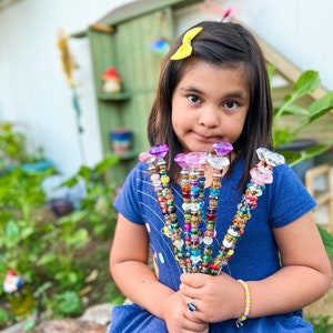May include: A young girl holds a bouquet of colorful beaded wands with crystal-shaped tops. The wands are decorated with various beads, including clear, gold, and rainbow-colored beads. The girl is wearing a blue dress and a yellow bow in her hair.