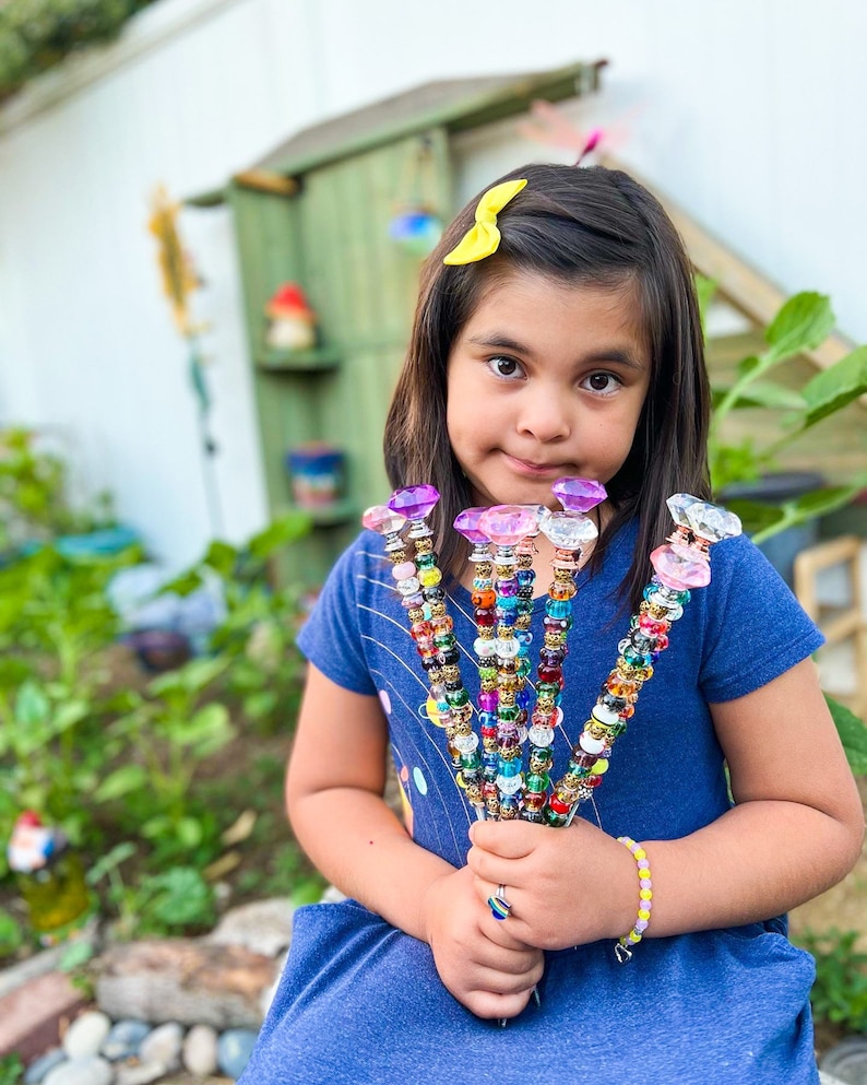 May include: A young girl holds a bouquet of colorful beaded wands with crystal-shaped tops. The wands are decorated with various beads, including clear, gold, and rainbow-colored beads. The girl is wearing a blue dress and a yellow bow in her hair.