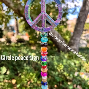 May include: A peace sign charm with a rainbow iridescent finish, attached to a beaded string with colorful beads. The beads are a mix of clear, turquoise, orange, red, green, and gold. The text "Etsy shop: elena monroe boutique" is visible in the image.