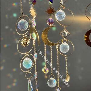 Peut inclure: Attrape-soleil c&eacute;lestes dor&eacute;s avec des accents de cristal transparent. Les attrape-soleil pr&eacute;sentent des formes de soleil et de lune, des sph&egrave;res de cristal et des pendentifs en cristal suspendus. Le nom de la boutique Etsy "elena monroe boutique" est visible.