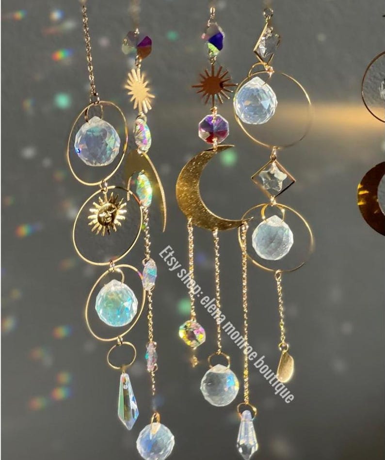 Peut inclure: Attrape-soleil c&eacute;lestes dor&eacute;s avec des accents de cristal transparent. Les attrape-soleil pr&eacute;sentent des formes de soleil et de lune, des sph&egrave;res de cristal et des pendentifs en cristal suspendus. Le nom de la boutique Etsy "elena monroe boutique" est visible.
