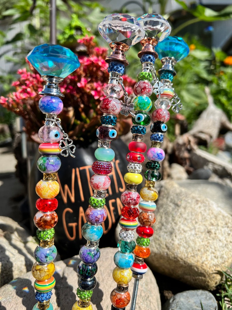 May include: Four colorful beaded garden stakes with crystal tops. Each stake features a variety of colorful beads, including clear, red, yellow, green, blue, and purple. The tops of the stakes are crystal-shaped and have a silver finish.