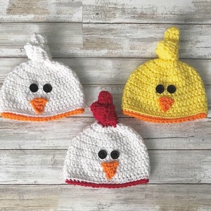 K&ouml;nnte beinhalten: Drei geh&auml;kelte M&uuml;tzen in Form von K&uuml;ken. Eine M&uuml;tze ist wei&szlig; mit einem orangefarbenen Schnabel und einem roten Bommel, eine ist gelb mit einem orangefarbenen Schnabel und einem gelben Bommel und eine ist wei&szlig; mit einem orangefarbenen Schnabel und einem roten Bommel.