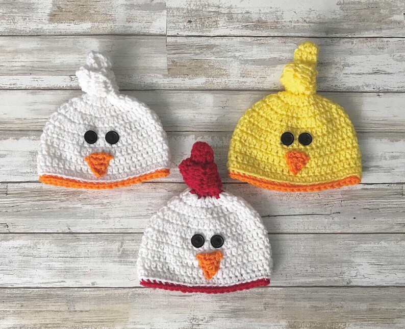 K&ouml;nnte beinhalten: Drei geh&auml;kelte M&uuml;tzen in Form von K&uuml;ken. Eine M&uuml;tze ist wei&szlig; mit einem orangefarbenen Schnabel und einem roten Bommel, eine ist gelb mit einem orangefarbenen Schnabel und einem gelben Bommel und eine ist wei&szlig; mit einem orangefarbenen Schnabel und einem roten Bommel.