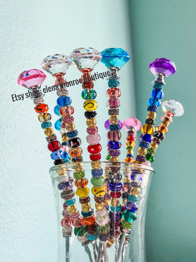 May include: A set of six colorful beaded sticks with crystal diamond-shaped tops. The sticks are arranged in a glass vase. The beads are various colors, including pink, red, blue, green, yellow, and orange.