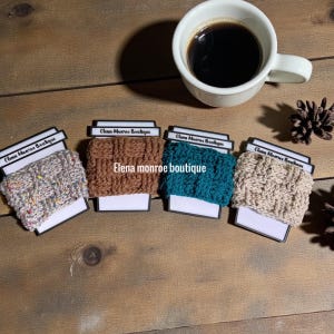 May include: Four knitted coffee cup sleeves in various colors: speckled gray, brown, teal, and beige. Each sleeve is displayed on a white card with the text "Elena Monroe Boutique". A white coffee cup filled with black coffee sits above.