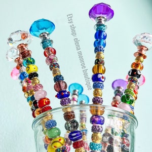 May include: A close-up of six colorful beaded wands with crystal tops. The wands are arranged in a glass vase. The beads are a mix of colors, including blue, green, red, yellow, and purple. The crystal tops are clear, blue, purple, and pink. Etsy shop: elena monroe boutique