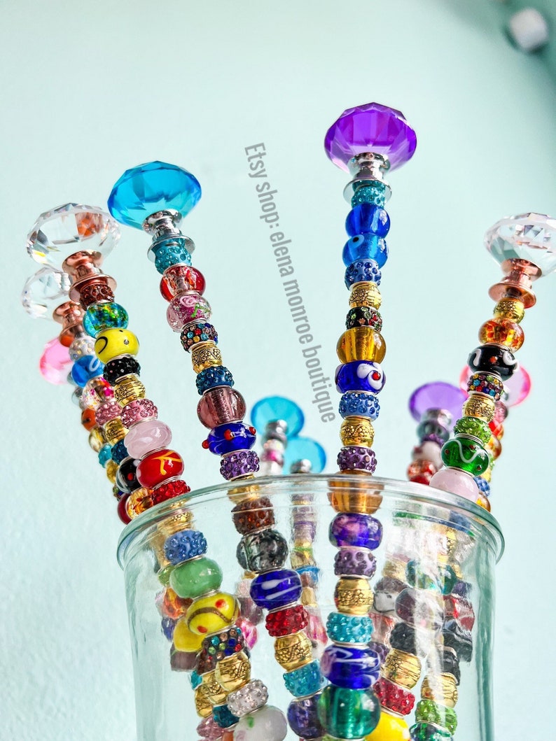 May include: A close-up of six colorful beaded wands with crystal tops. The wands are arranged in a glass vase. The beads are a mix of colors, including blue, green, red, yellow, and purple. The crystal tops are clear, blue, purple, and pink. Etsy shop: elena monroe boutique